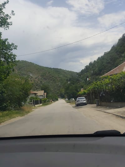 The image depicts a street scene in Jeleznitsa, Bulgaria, as viewed from inside a car. The road stretches into the distance, flanked by houses and lush greenery. Mountains rise on either side of the road. A silver car is parked on the right side of the road. A Bulgarian flag flutters near one of the houses. The sky is overcast with cloudy. The car's dashboard and windshield wiper are visible at the bottom of the frame.