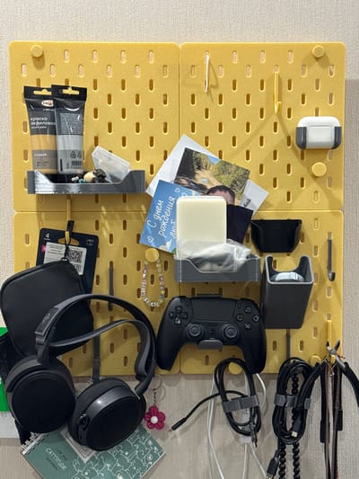 An overhead shot shows a yellow pegboard wall with various items neatly organized and hung. On the top left, two tubes of acrylic paint are attached to the board, one labeled "краска акриловая" (acrylic paint) and the other "студия" (studio). To the right of the paint tubes, a small gray shelf holds a clear plastic container with what appears to be small figurines and beads. Above the shelf, a white wireless earbud case is mounted to the board.

In the center of the pegboard, a few pieces of paper are tucked into the holes, including a photograph of a natural landscape and a birthday card with Russian text. To the right of the papers, a white bar of soap is placed in a clear plastic bag, resting on a gray tray. A small black container is hung to the right of the soap, and below it, another gray container holds a light blue object.

Below the shelf, a black gaming controller is mounted horizontally on the pegboard. To the left of the controller, a pair of black headphones with a gray headband hangs. Below the headphones, a green sketchbook with hand-drawn doodles and text reading "I LIKE YOUR CATTITUDE SKETCHBOOK" is placed.

Various cables, including USB cables and charging cords, are bundled and hung on the right side of the pegboard. A small pink flower-shaped keychain is attached to the headphones. A small set of pliers with red handles hangs on the far right of the pegboard. The pegboard is mounted on a textured, beige wall, suggesting an indoor domestic environment. There is no indication of the location being Ulan-Ude, Russia.