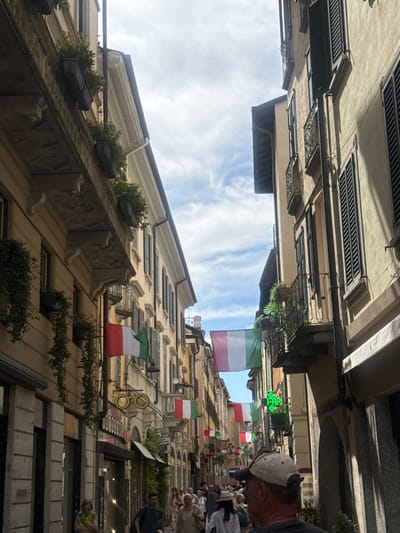 The image shows a narrow street in Como, Italy, lined with buildings on both sides. Italian flags are prominently displayed, hung across the street and attached to buildings. Several people are walking down the street, some are wearing hats and casual clothing. The buildings appear to be old, with balconies and shutters on the windows. There are shops and businesses visible at street level, indicated by signage. A bright green illuminated "FAR" sign, likely for a pharmacy, is visible on the right side of the street. The sky is cloudy with patches of blue. The overall scene depicts a lively street in a European city.