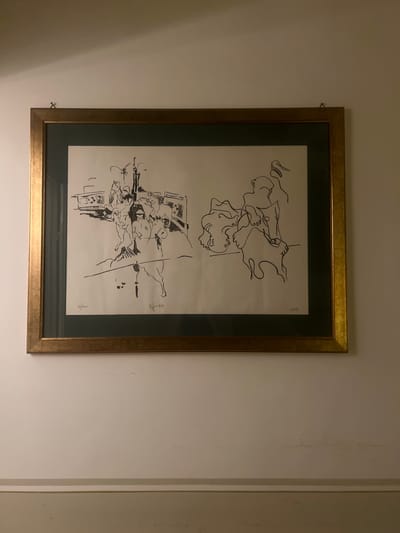 A framed black and white drawing hangs on a white wall. The frame is gold-colored. The drawing depicts two scenes. The top scene shows abstract figures, possibly people. The bottom scene shows a more detailed setting, possibly an interior with figures and objects such as furniture. A signature and a notation such as "9/100" appear in the lower right corner of the drawing. The scene lacks contextual information to place it in Siena, Italy.