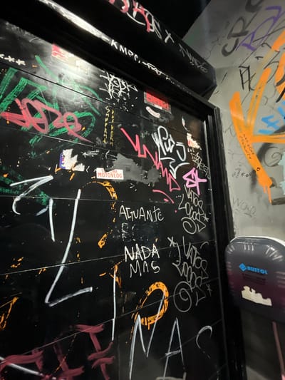 This image shows a wall or door covered in graffiti. The writing includes phrases such as "AGUANTE," "NADA MAS," "WOD," and "Lomas de Zamora." There's also a sticker that reads "MOTOVLOG," and the brand "BUSTOL" is visible near the top. The wall or door is black, with colorful graffiti in orange, white, green, pink, and purple. The photograph is taken from a low angle, looking up at the surface. The location, as indicated by the graffiti, is Lomas de Zamora, Argentina.