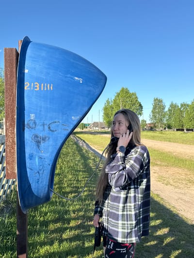The image depicts a young woman utilizing a weathered blue public telephone booth in a rural setting, identified as Ust'-Silayka, Russia. The woman, with long light-brown hair and a fair complexion, is shown in profile from the chest up, holding a black telephone receiver to her right ear with both hands. Her gaze is directed towards the right, away from the camera, conveying an attentive or pensive expression. She wears a plaid flannel shirt in shades of blue, green, and white, with the visible lower portion of dark shorts or pants featuring a repeating pattern of red chili peppers and white text, partially legible as "Salz und Pfeffer" (German for "Salt and Pepper"). A black smartwatch with a pink band is visible on her left wrist, displaying "7:37".

The telephone booth is a distinct, curved blue structure mounted on a brown, rusted metal post. Yellow numbers "2131111" are painted on its upper-left surface, along with various dark, scratched graffiti marks below them, including circular shapes and what appears to be a stylized "O+C=" and a heart. The black phone handset is connected to the booth by a silver coiled cord.

The environment is characterized by a bright, clear blue sky overhead. The ground in the foreground and middle ground consists of green grass with patches of exposed dirt. A dirt road or path extends into the background, curving towards the right. Several trees with vibrant green foliage, possibly birch trees, are scattered across the middle and background. In the distance, multiple rustic wooden houses or buildings are visible, indicative of a village. A wooden fence is partially seen to the left of the telephone booth, and a utility pole with wires stands along the dirt road. The scene is illuminated by strong natural sunlight, casting distinct shadows on the grass to the right.