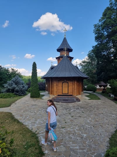 A young woman with long brown hair is standing on a stone pathway next to a wooden church. She is wearing a white t-shirt, blue denim shorts, and white sneakers. She is carrying a backpack with blue, pink, and white patterns. The church is a traditional Orthodox wooden structure with a dark grey, multi-tiered roof topped with golden crosses. The church is situated in a lush green setting with trees and bushes. The sky is bright blue with scattered white clouds, indicating it is daytime. The weather appears to be pleasant and sunny. The scene is likely outdoors at a religious site. The image was taken in Hagimus, Moldova. There is no visible text in the image.