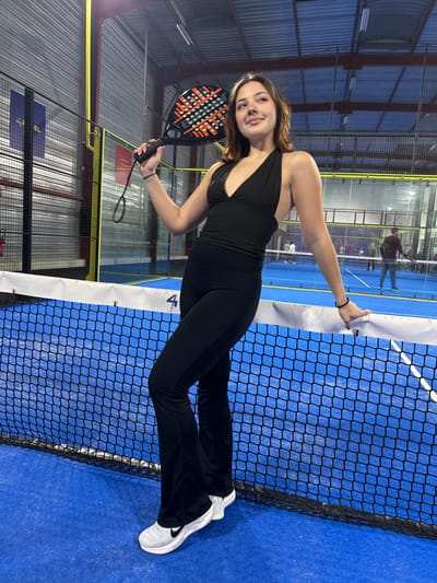 An indoor portrait displays a young woman standing upright on a blue padel tennis court, smiling directly at the viewer. She is positioned next to the net, which spans horizontally across the image. The woman wears a black halter-neck sports top, black flared athletic pants, and white athletic shoes with a black Nike swoosh logo. In her right hand, she holds a black and orange/red padel racket, displaying the brand "SIUX" on its surface. Her left hand rests casually on the white band of the padel net.
The padel court features transparent glass walls, typical of the sport, through which blurred figures of other people are visible in the background, suggesting an active sports complex environment. The ceiling reveals industrial-style lighting fixtures, indicating an indoor setting, likely in the evening or at night. Metal support structures for the court enclosure are also visible. On the white band of the net, a number "4" is partially discernible, possibly indicating the court number. Further in the background, fragmented text "FEL" can be seen on a wall or banner. The scene captures a moment of leisure or post-activity on a padel court in Eckbolsheim, France.
