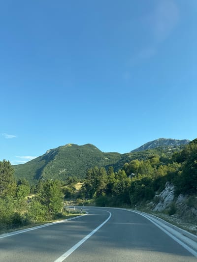 A winding asphalt road curves through a mountainous landscape under a clear blue sky. The road has white lane markings. To the left, lush green trees and vegetation cover a steep hillside. In the distance, a large, verdant mountain rises, its peak dotted with patches of green grass. To the right of the road, the terrain is rocky and covered with trees and shrubs. A few white posts with reflective markers line the side of the road. The scene is set in the city of Kopito, Montenegro.