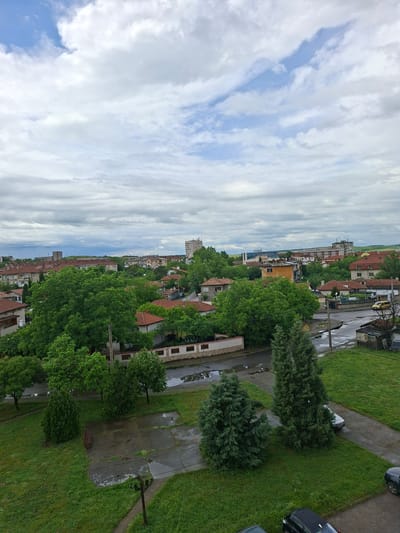 The image depicts an elevated view of a section within the city of Galabovo, Bulgaria. The sky occupies the left portion of the frame, displaying a partly cloudy condition with blue patches visible. The right portion features a residential area with a street running along the middle, lined with buildings, trees, and parked vehicles. The architecture consists of low to mid-rise buildings, many with red-tiled roofs. The environment includes greenery such as grass, trees and bushes. Concrete sidewalks are visible alongside roadways. No people are directly observable in the image.