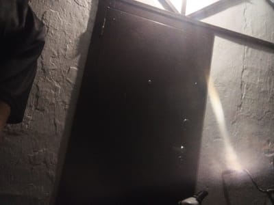 This media file shows a dimly lit, possibly industrial or basement setting. The main subject is a large, dark, metallic object, likely a door or a safe, with visible hinges on the left side. The wall behind it is rough and textured, painted a light gray. To the left of the metallic object, a dark, blurry shape suggests a person wearing dark clothing, possibly a jacket or coat. In the lower right corner, a bright light source is visible, emitting a beam of light that illuminates the dusty air, suggesting it might be a flashlight or a spotlight. The overall atmosphere is gritty and somewhat mysterious, with a hint of abandonment or disuse. The lighting suggests it could be nighttime or an indoor location without natural light. There is no visible text in the image. The location context provided is Leontyevskoye, Russia.