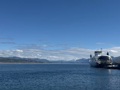 The image shows a ferry docked at a pier in Lødingen, Norway. The ferry, which is primarily white and black, is positioned on the right side of the frame, facing towards the left. It has a prominent upper structure with windows and what appears to be a bridge. The ferry's open bow ramp suggests it is ready for loading or unloading. The water in front of the ferry is a deep blue and appears calm with small ripples.
In the background, across the water, a range of mountains is visible. Some of the peaks are snow-capped, indicating a mountainous terrain likely characteristic of the Lofoten Islands region, where Lødingen is situated. The sky is a clear, vibrant blue with scattered white clouds, particularly concentrated near the horizon above the mountains. The overall lighting suggests it is daytime, with the sun casting gentle light on the scene.
The pier area to the right of the ferry shows some industrial elements, including what looks like a building or storage facility, along with some equipment and ropes. There are no people visible in the image, nor are there any other vessels in the immediate vicinity besides the docked ferry. The scene conveys a sense of a functional port in a scenic, coastal environment.