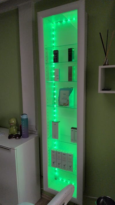 The image shows a lit display shelf with several small boxes. The shelf is illuminated by green LED lights. The boxes appear to be cosmetic or skincare products, with some visible text that is difficult to decipher. To the left of the shelf, there are some dark-colored sticks. Above the shelf, there is a small white shelf with a black object on it. Below the display shelf, a green wall is visible. To the right of the display shelf, part of a white cylindrical object is visible. Further down on the right, there is a white cabinet with a blue and purple water bottle and a pair of sunglasses on top. The image appears to be taken indoors, possibly in a home or retail setting. The lighting suggests it could be evening or a dimly lit room.