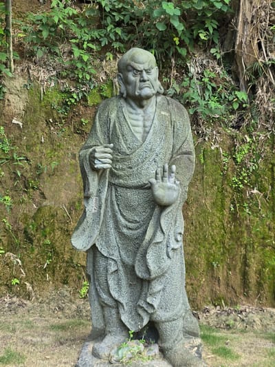 A stone statue of a formidable, bald-headed man with a stern expression stands in front of a moss-covered, rocky hillside with dense green foliage. The statue is depicted wearing a draped robe. The man's right hand is raised with fingers splayed, while his left hand holds a small object. The ground in front of the statue is covered with dry grass and a few small green shoots. The overall scene suggests an outdoor setting, possibly in a temple or garden, located in Xiaoshan District, China.