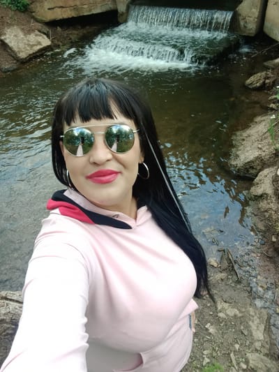A woman is taking a selfie near a waterfall. The woman has long, black hair, wears mirrored aviator sunglasses and bright pink lipstick. She is wearing a light pink hooded sweatshirt with contrasting black and red trim around the hood. She is standing on a rocky bank next to a body of water that appears to be a stream with a small waterfall in the background. The surrounding environment consists of rocks, dirt, and some greenery. The image is taken from a high angle, looking down at the woman and the scene.