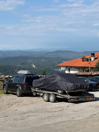 An exterior, low-angle, medium shot shows a dark gray SUV attached to a trailer carrying a car covered by a black tarp. The SUV has a gray roof box and is parked on a gravel surface. The trailer has a license plate that reads "B 3555 EH". In the background, a red-tiled roof house with white walls and a chimney is visible. Beyond the house, a hilly landscape with layers of mountains covered in green trees and some buildings is seen under a blue sky with scattered clouds. The scene appears to be in a rural area, possibly on the outskirts of Arbanassi, Bulgaria, given the architectural style of the house and the mountainous terrain. There are no people visible in the image.