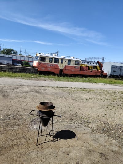 The image depicts an outdoor industrial scene on a sunny day in Shimanovsk, Russia. In the foreground, a rusted metal braizer stands on a black metal tripod. The ground is a mixture of dirt, gravel, and cracked concrete. In the background, a train is parked on tracks. The train is composed of a yellow and red locomotive and several cars. One car is a crane, and another is a white container car with Cyrillic lettering that reads "МОДУЛЬ". The train appears to be stationary. The sky is clear blue with a few wispy clouds. The overall scene suggests a maintenance or storage yard for railway equipment.