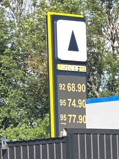 The image shows a gas station sign in Shatava, Ukraine. The sign has a white square with a black triangle in the center, and the words "UKRNAFTA" in yellow letters below it. The sign also displays fuel prices: 92 octane at 68.90, 95 octane at 74.90, and another 95 octane at 77.90. The sign is tall and slender, with a yellow border. The background consists of green trees and a blue sky. A dark fence with vertical slats runs along the bottom of the image.
