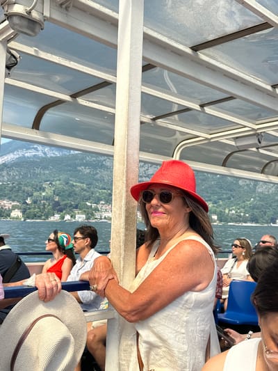 This is a candid, close-up shot of a woman wearing a red hat and sunglasses, leaning on a railing while on a boat. She is smiling at the camera, her expression suggesting a relaxed and happy mood. Behind her, other passengers are visible, some seated and looking out at the scenery. The backdrop is a picturesque view of a lake with mountains and a village on the shoreline, characteristic of the Italian Lake Como region, specifically Griante. The sunlight suggests it's daytime, likely late afternoon given the warm, golden light. The woman is wearing a white dress, and her arm is adorned with rings, one of which is a notable gold band. The overall atmosphere is one of leisure and enjoyment, perhaps a scenic tour on the lake. The boat itself appears to be a ferry or tour boat, with its metal framing and seating visible.