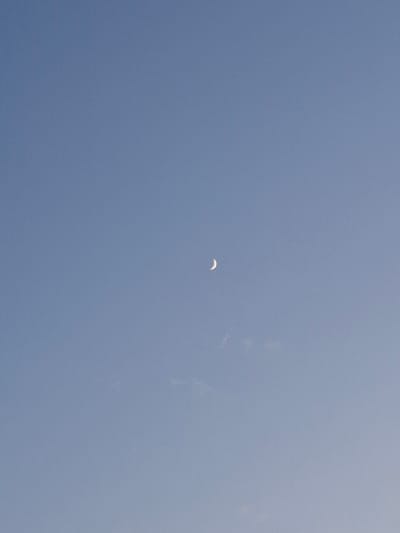 A clear, pale blue sky dominates the image. A sliver of the crescent moon is visible in the upper right quadrant of the frame, appearing as a thin, bright curve against the expanse of the sky. There are no other discernible objects, people, or structures in the image. The environment is solely the sky.