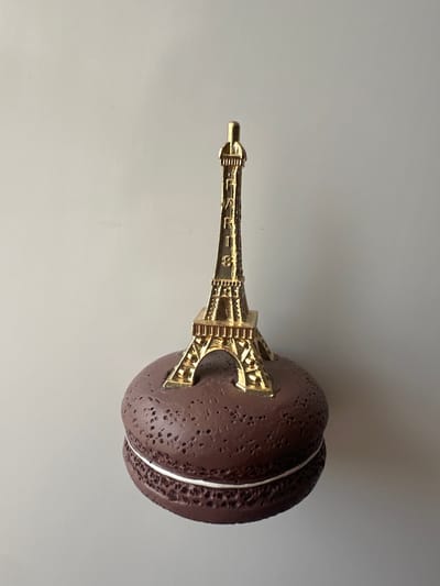 The image shows a close-up, horizontal shot of a novelty item resembling a dark brown macaron with a miniature golden Eiffel Tower protruding from its center. The macaron has a textured surface with many small indentations, and a thin white band encircles its middle, separating the two halves. The Eiffel Tower is detailed, featuring intricate lattice work and architectural elements. The object is placed on a plain, light gray surface, likely a table or floor, which takes up the majority of the frame. The lighting is neutral, suggesting an indoor setting with artificial light. No people are visible, and the scene depicts a static object rather than an activity or event. The location context provided is Marten, Bulgaria, but there are no visual cues within the image itself to confirm this specific location. There is no visible text in the image.