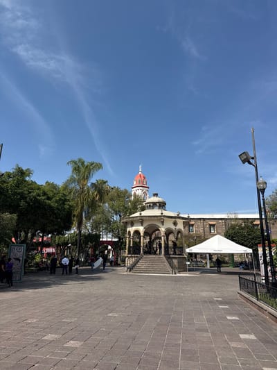 A scene features a tiled walkway and a partly visible building with arched structures and stairs leading up to it. The building is topped with a dome that has a red roof. There's a white tent-like structure beside the building. A street light with a spotlight faces towards the right of the image. In the background, green trees contrast with a bright blue sky dotted with wispy clouds. People are present near the walkway.