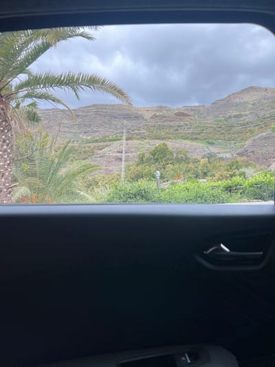 The image depicts a landscape seen through a car window, likely taken on a cloudy day in Mogán, Spain. The foreground contains a blurry view of the car's interior including a car vent and door handle. The landscape features a terraced mountain covered in vegetation, including palm trees and other greenery. A tower is visible midway up the mountain. The sky is overcast with grey clouds.