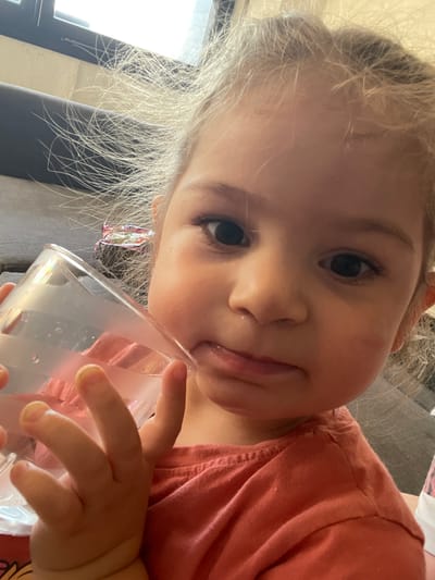 A young child with light hair and fair skin is drinking from a clear, decorated glass. She wears a salmon-colored shirt. The setting appears to be indoors, possibly a home in Senden, Germany, suggested by the original context (though not directly visible in the image itself). The background includes a gray sofa and a window.