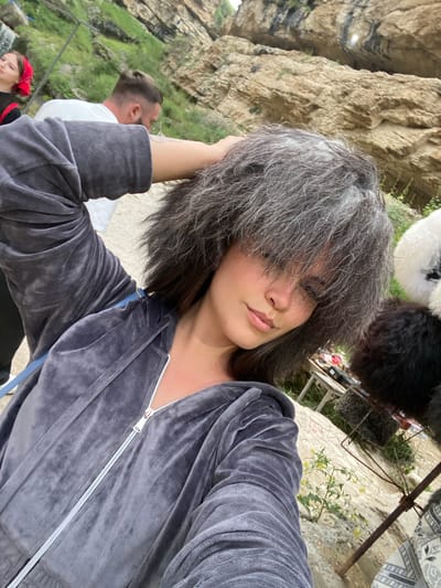 A selfie is taken by a young woman with a grey, fluffy wig and a grey velour tracksuit, with her right arm raised and hand on her head. She is in the foreground with a blurry background of a rocky landscape and some people. In the background, a man with a bun hairstyle is visible. There are also some stalls with hats. The location appears to be the village of Saltá in the Republic of Dagestan, Russia, known for its ancient stone towers and picturesque mountain scenery.