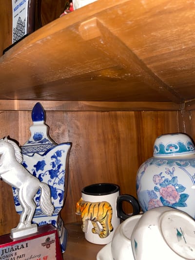 The image shows a close-up view of items displayed on a wooden shelf. On the left, a silver horse figurine stands on a red and silver plaque. Behind the horse is a tall, white ceramic vase decorated with blue floral patterns. To the right of the vase is a white mug with a black handle, adorned with a painted tiger design. Further to the right, a large, round white ceramic jar with a lid features blue and pink floral motifs. In the foreground, the bottom of two white ceramic bowls or dishes is visible, with one showing a faint green logo. The wooden shelf is visible above and below the objects, with a textured grain pattern. The overall impression is of a collection of decorative items on display in a home or office setting. The location is identified as Margorejo, Indonesia, suggesting this collection is situated within that region.