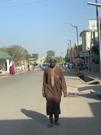 The image depicts a street scene in Hotoro South, Nigeria, under a clear, bright sky.
In the immediate foreground, an adult male with dark skin walks forward, slightly right of the center. He wears a long-sleeved, brown traditional tunic with light-colored embroidery around the neck and chest, a patterned dark skullcap, and dark sandals. He looks directly at the camera, casting a long shadow to his left.
The street, appearing to be asphalt or packed earth, extends into the background, where other pedestrians and vehicles are visible. On the left side of the street, a person in a red garment walks away from the camera near a building with a light green pitched roof. Further down, more individuals and trees with sparse foliage are seen.
On the right side of the street, a row of buildings lines the road, including "MUKADDAM MEDICAL STORE" with visible contact information (08032797073). Modern streetlights with solar panels are installed along this side. A rectangular sign featuring a portrait of a man labeled "SARAYYA" and "HON. NASIR ALIYU AHMED" is attached to a streetlight pole. A street vendor's cart with a striped umbrella, displaying items, is positioned near the sidewalk. An open concrete drainage ditch runs parallel to the street on the right. Several other individuals are present on the sidewalks and near the vendor stalls. A silver car is visible in the middle distance on the road.