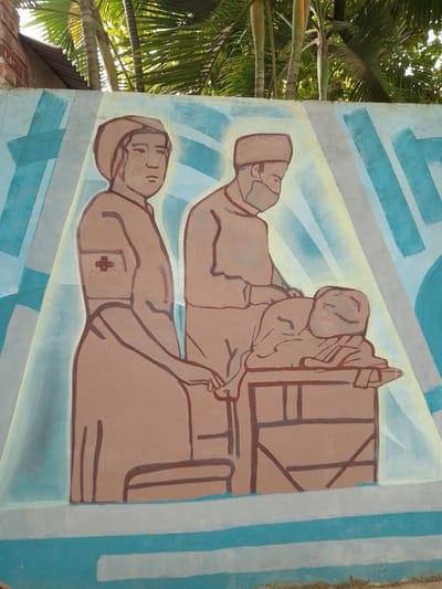 The image is a close-up shot of a mural painted on a wall in Villa Hayes, Paraguay. The mural depicts a medical scene, likely representing healthcare or historical medical practices.

In the mural, there are three figures. On the left is a female figure in a long dress with a red cross visible on her sleeve, indicating a nurse or medical worker. To her right is a figure dressed in what appears to be surgical attire including a cap and mask; likely a surgeon. They both appear to be attending to a third figure, who is lying on what appears to be an operating table.

The style of the painting is somewhat simplistic, with thick outlines. The color palette is limited, with shades of brown, blue, white, and yellow predominating. The background consists of geometric shapes in shades of blue, white, and yellow. Above the mural, there is green foliage, suggesting the wall is in an outdoor location possibly a garden or park. The wall is painted with horizontal blue and white stripes above the base.