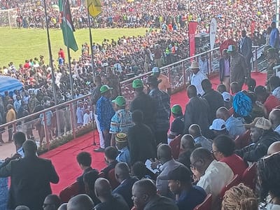 This media file depicts a bustling outdoor public gathering, likely a rally or official ceremony, held in a stadium or large open ground in Siaya, Kenya. The scene is characterized by a massive crowd of people, with individuals filling both the foreground seating areas and extending into the distant stands and a grassy field.
In the middle ground, a prominent red-carpeted platform serves as a stage or VIP section, enclosed by metal railings. On this platform, numerous individuals are visible, including several men wearing light-colored fedoras or matching green caps, some in dark suits, and others in military-style camouflage uniforms, suggesting the presence of officials, dignitaries, and security personnel. A microphone stand is positioned on the red carpet, indicating that speeches or addresses are being given or are imminent. A few children, one also wearing a green cap, are present in this area.
Above the platform, several flags are flying, clearly showing the Kenyan national flag (green, red, black, and white), alongside another green flag and a yellow flag. To the right of the stage area, a white banner featuring the acronym "NCAA" is partially visible, along with a red banner.
The event takes place during the daytime under clear skies, with bright natural light illuminating the large assembly. The overall impression is one of a significant public event drawing a substantial crowd, with attendees focused towards the stage area.