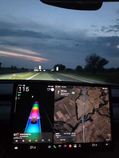 The image depicts the interior of a Tesla vehicle driving on a highway at dusk or early night, specifically on the A4 motorway in Bulgaria, within the general vicinity of Dositeevo, en route to Dimitrovgrad.
The prominent central infotainment screen displays a digital dashboard on the left, an autopilot visualization in the center, and a navigation map on the right. The vehicle is traveling at 115 km/h, indicated by the large numeral, with the gear selector in 'D' (Drive) and low beam headlights active. The battery charge is at 68%, and the ambient temperature is 24°C, with the time noted as 21:18. The autopilot visualization shows the car centered on a multi-colored road. Beneath this visualization, the music player displays "IN DA STREETZZ" by "SHMEKERA" as the current track.
The navigation system on the right indicates "Dimitrovgrad" as a key point, with 38 km remaining. The estimated time of arrival is 23:52, with 2 hours and 34 minutes of driving remaining to cover 187 km to the final destination, Kilifarevo, at which point the battery is projected to be at 15%. The map displays an aerial view of agricultural fields traversed by the A4 motorway. A "PASSENGER AIRBAG ON" indicator is visible, along with a compass pointing "NW". The cabin climate control is set to 21.5 degrees Celsius.
Through the windshield, the highway extends forward into the twilight, illuminated by the vehicle's headlights and distant, blurred lights from other cars and sparse streetlights. Red roadside markers or reflective elements are visible along the left side of the highway. The sky transitions from a dark blue overhead to lighter grey and faint orange-pink hues near the horizon, indicating the recent setting of the sun. The surrounding landscape consists of indistinct, dark trees and broad, flat agricultural fields. The windshield itself shows minor surface debris. No individuals are visible in the image.