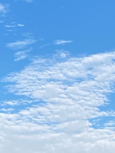 A bright blue sky is filled with fluffy white clouds. A pale, crescent moon is visible in the upper center of the frame, partially obscured by clouds. The image appears to be a photograph taken from ground level, looking upwards towards the sky. There are no people, buildings, or other man-made objects visible in the frame. The scene is serene and tranquil, with the clouds creating a sense of depth and movement. There is no information within the image to identify the specific location within the city of Unwana, Nigeria.