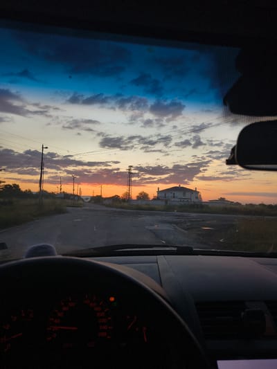 The image is taken from inside a car, looking out through the windshield. The scene depicts a road leading towards a rural area with a large white house visible in the distance. The sky is filled with dramatic clouds, illuminated by the warm hues of a sunset, with shades of orange, pink, and blue. Utility poles and wires crisscross the sky, indicating the presence of infrastructure. The road appears to be asphalt and shows signs of wear and tear, with some puddles of water. The dashboard of the car is partially visible in the foreground, with illuminated gauges indicating it is either dusk or the car is in motion. The location is identified as the city of Padarsko, Bulgaria, suggesting a view from a car parked or driving on the outskirts of this settlement.