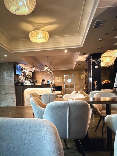 The image shows the interior of a modern cafe, possibly located in Novinki, Russia. The cafe features a well-lit, contemporary design with a bar area, seating arrangements, and decorative elements.
In the foreground, several plush, light-colored armchairs are visible, suggesting a comfortable dining or lounging experience. The chairs have a textured fabric and a modern, streamlined design. One chair in the center is particularly prominent, showcasing its cushioned backrest with a vertical seam detail.
The middle ground reveals a bar area with a marble-effect counter and a backlit surface. Behind the counter, shelves stocked with bottles and glasses are illuminated, indicating a bar setup. Two individuals appear to be working behind the bar, one of whom is partially visible. A television screen mounted on the wall displays an image.
The background includes a series of pendant lights with an intricate, textured design, casting a warm glow. The ceiling is adorned with molding, and ventilation grilles are visible. Further back, there are doors with circular signage and decorative elements on a dark pillar. The flooring appears to be a polished wooden surface, reflecting the ambient light. The overall ambiance is one of a sophisticated and inviting establishment.