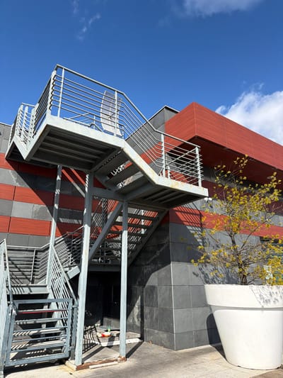 The image captures an exterior view of a modern building in an urban area of Viseu, Portugal, under a clear blue sky with scattered white clouds. A multi-flight external metal staircase, constructed from galvanized steel with horizontal bar railings and open risers, ascends against the building's facade on the left. The building's lower section is clad in dark grey horizontal panels, interspersed with narrower horizontal bands of reddish-orange material. An upper section of the building exhibits a larger expanse of reddish-orange cladding with sharp architectural lines. Direct sunlight casts distinct shadows from the staircase onto the building facade and the paved ground below. Beneath the lowest flight of stairs, near a dark recessed doorway, a white plastic bottle with a red cap and other small discarded items are visible on the ground. To the right of the staircase, a small, deciduous tree with sparse, nascent yellow-green leaves is planted in a large, white, conical planter pot. A white security camera is affixed to a reddish-orange band on the building facade, positioned above the tree. No individuals are present in the scene, and no overt actions or interactions are depicted.