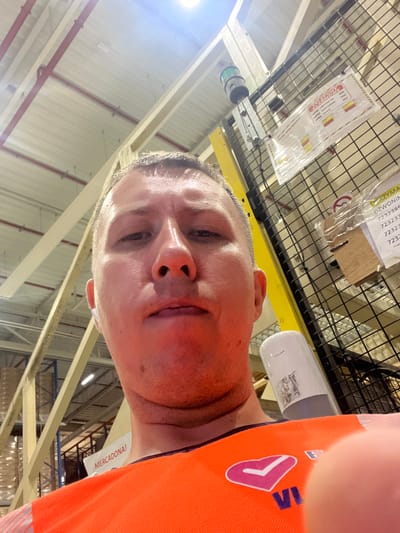In a wide-angle, low-angle shot, a man wearing an orange high-visibility vest with a pink heart and checkmark logo is positioned in the foreground, his face filling the center of the frame. He is looking directly into the camera with a neutral expression. He has short, light brown hair and appears to be wearing a white wireless earbud in his left ear.
The background reveals a large industrial indoor space, likely a warehouse. Above, a bright light source illuminates the scene. Exposed ceiling beams, some painted red and others light wood, crisscross overhead. To the right, a black wire mesh fence is visible, with various labels and signs attached. One sign, in Polish, appears to be a safety or operational instruction, with text such as "MILIONY ALARMOWE" and numbers. Another sign partially visible reads "DZWONIN..." and a series of numbers. Further in the background, tall shelving units filled with stacked items are discernible, suggesting a storage facility. A yellow vertical support beam is also present on the right side.
A partially obscured white cylindrical object, possibly a dispenser, is attached to the mesh fence behind the man's shoulder. A white paper with the text "MERCADONA!" is visible on the man's left side, tucked into his vest. The overall environment suggests a logistical or distribution center. The image appears to have been taken in the city of Oława, Poland, given the presence of Polish text on the signs.