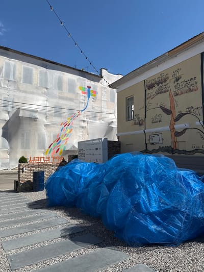 The image depicts an outdoor scene in the city of Yelets, Russia, likely a public space or courtyard. In the background, a building is covered in white protective sheeting, with a colorful mural of a dragonfly and a trail of colorful dots painted on it. To the right, another building features a mural with stylized drawings of plants and figures, rendered in shades of yellow and brown. A light string with bulbs stretches across the top of the scene. In the foreground, a pile of large blue netting lies on a gravel surface interspersed with rectangular paving stones. A brick structure with a sign that reads "АРТ ЕЛЕЦ" (ART YELETS) is visible to the left of the netting, accompanied by two blue barrels and a small evergreen plant. The sky is clear and blue, with a faint crescent moon visible. No people are present in the image.