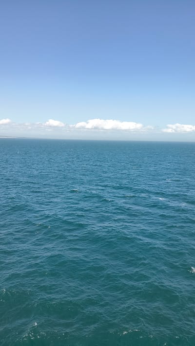 The image shows a vast expanse of the ocean under a clear blue sky. The water is a deep teal color with small waves rippling across its surface, reflecting sunlight. In the distance, a faint line of clouds is visible on the horizon, possibly marking the edge of land. The scene is serene and open, conveying a sense of immensity and tranquility. No people, specific structures, or identifiable landmarks are present. The location is not specified.