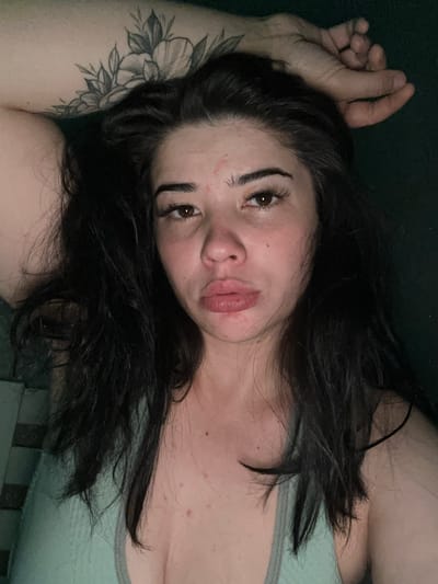 A close-up, high-angle, dimly lit, medium-shot photograph shows a young woman with long, dark, wavy hair lying down. Her head is tilted back, and her right arm is bent and resting behind her head, with her hand touching her forehead. A floral tattoo is visible on her upper right arm. She has dark brown eyes, well-defined eyebrows, and full lips. Her skin appears fair with some redness and visible pores, particularly on her cheeks and forehead. She is wearing a light blue-green tank top. The background is dark green and out of focus. The image does not provide any information about the city of Gavrilov Posad, Russia, or any other specific location.