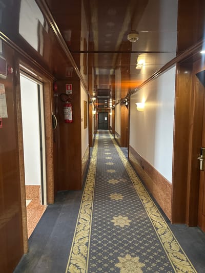 The image shows a hotel corridor in San Martino Buon Albergo, Italy. The corridor has dark wood paneling on the walls and ceiling, which have a high gloss finish. The floor is covered with a dark blue carpet featuring a repeating pattern of small square motifs and larger starburst designs, bordered by a decorative yellow pattern. On the left side of the corridor, there are several closed doors and an open doorway revealing another area with a different floor pattern. A red fire extinguisher is mounted on the wall near the entrance. The right side of the corridor features more closed doors and wall-mounted sconce lights that cast a warm glow. The corridor recedes into the distance, creating a sense of depth.