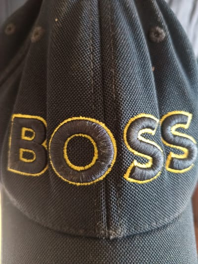 The image shows a close-up of a black baseball cap with the word "BOSS" embroidered in black with a yellow outline. The cap is made of a textured fabric. The lighting is somewhat harsh, creating shadows that highlight the texture of the fabric and the embroidery. There are no people or other identifiable objects in the frame. The image does not provide any information about the location in Huntingdonshire, United Kingdom.