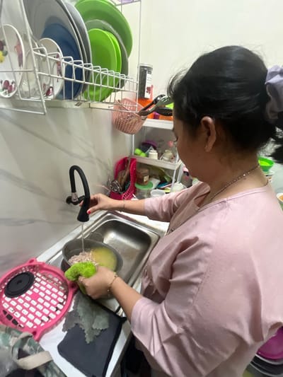 A woman with dark hair pulled back is in a kitchen, likely in Serdang Kulon, Indonesia, preparing food. She is wearing a pink, striped long-sleeved shirt and a silver necklace. Her right hand, adorned with rings, is holding a green sponge, and she is using it to clean the contents of a pot filled with food and liquid, which is situated in a stainless steel sink. The faucet above the sink is turned on, with water running into the pot. To the left of the sink is a pink plastic strainer with a black insert. In the background, a dish rack holds various plates and bowls, and shelves are stocked with kitchen items and containers. The wall behind the sink has a marble-like pattern.