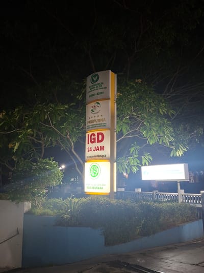 A nighttime, eye-level exterior shot shows a tall, illuminated signpost in Lampeuneurutblang, Indonesia, in front of a hospital. The signpost has multiple panels with text and logos. The top panel displays the logo of "RSUD MEURAXA BANDA ACEH" and a phone number "0651-43097". Below that, a panel indicates "PARIPURNA" with a star rating. The largest panel prominently features "IGD" in red letters and "24 JAM" below it, along with a website address "http://rsum.bandaacehkota.go.id/". The lowest visible panel shows the logo of the Indonesian Ulema Council (MUI) and "RSUD MEURAXA".

Behind the signpost, lush green trees with dark branches are visible against a dark sky. In the foreground, there is a blue-tiled planter filled with various green plants, including shrubs and tall grass. A bright white illuminated sign is visible to the right of the main signpost, but its text is partially obscured by glare. The overall scene suggests the entrance to a hospital at night, with clear signage for its services.