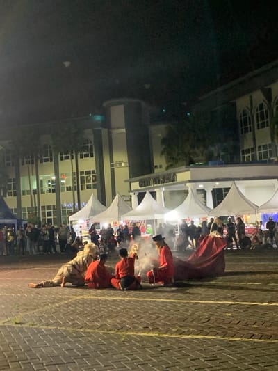 The image shows a performance in a large outdoor space in front of a multi-story building in Kecamatan Batu, Indonesia. The building is lit up at night. There are several white tents set up in front of the building, and a large group of people are gathered in the area, watching the performance. There are at least 4 performers in the center of the image, they are sitting or crouching. The performers are wearing red clothing. The performer on the left is in a crouched position holding a white cloth and in the foreground there is a white and light brown object that appears to be a costume. A large red cloth is spread across the pavement, and a large amount of smoke is billowing up from the center of the image. There are several more people standing in the background around the white tents. The floor is made of brick. The image is taken at night and is well lit. The scene is set in front of the building, which is located at latitude -7.867 and longitude 112.514.