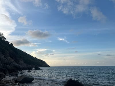 The image captures a coastal scene in Baan Chaloklum, Thailand, featuring a rocky shoreline meeting the ocean under a partly cloudy sky. A landmass, possibly a hillside or a headland, covered in vegetation, occupies the left side of the frame, with exposed rocks along its base. The sea extends to the horizon with gentle waves lapping the shore. Several large rocks are scattered along the beach in the foreground. The sky is a mix of blue and white, with scattered clouds.