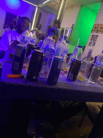 A bar scene displaying several opened bottles and cans on a table. Water bottles, a liquor bottle, and energy drink cans are visible, each pierced with a straw. An orange lighter sits near the table's edge. Patrons are faintly visible in the background. The scene is illuminated with vibrant blue and green lighting, creating a lively atmosphere.
