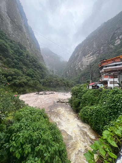 A muddy river rushes through a steep, lush valley. Buildings are precariously perched on the edge of the cliff overlooking the torrent. Mist and rain obscure the tops of the mountains that loom on either side. The vegetation is abundant, with green bushes and trees crowding the landscape. A few utility lines are visible across the valley. The overall atmosphere is wet and rugged.