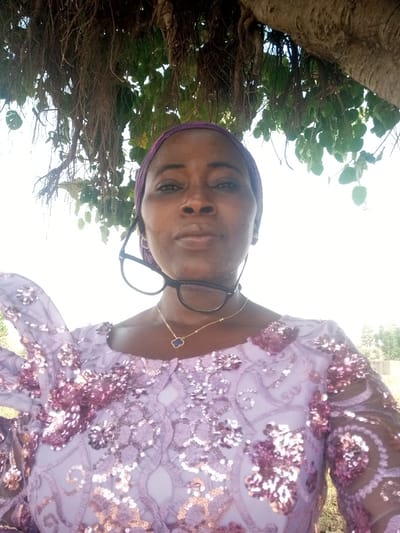 The image presents a headshot and upper body portrait of a middle-aged Black woman, captured outdoors in Gudige Sabo, Nigeria. The woman faces the camera directly with a neutral expression. She wears a purple fabric head wrap covering her hair. Black-framed eyeglasses are suspended around her neck, resting on her upper chest. A delicate gold-colored chain necklace with a small, dark blue, four-leaf clover pendant adorns her neck. She is dressed in a light purple garment, likely a top or dress, featuring a scoop neckline and extensive darker purple/magenta floral embroidery with iridescent sequins covering the visible chest, shoulders, and sleeves.

The background directly behind and above her features a large tree canopy. Prominently visible are numerous thin, dark aerial roots or vines hanging downwards, along with green foliage and a thick tree trunk or branch on the upper right. Glimpses of a bright, light-colored sky are visible through the tree's branches. In the distant background to the right, a blurred, light-colored structure and sparse vegetation can be vaguely discerned.