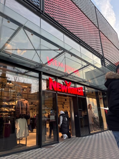The image captures the exterior of a "NEW YORKER" retail store located in Reichstett, France. The store features a prominent, illuminated red "NEW YORKER" sign above its main entrance. A clear glass canopy, supported by metal framework, extends over the entrance area. Above the glass storefront, the building's facade is clad in a textured, geometric pattern composed of red, pink, and dark gray/blue square or rectangular elements.

Through the left display window, various apparel is visible, including a brown hoodie and lighter-colored sweaters on mannequins, alongside sneakers displayed on shelves. The store's main entrance consists of open glass doors. A person wearing a white hoodie and a dark puffer vest is actively entering the store, with additional indistinct figures visible further inside. To the immediate right, the back of an individual in a dark parka with a fur-lined hood and blue jeans is partially visible on the paved sidewalk, facing away from the store. The sidewalk is constructed from interlocking light and dark gray bricks arranged in a checkered pattern. The upper right portion of the image shows a bright sky with some cloud cover. Reflections of trees and sky are present on the glass surfaces of the storefront and canopy.