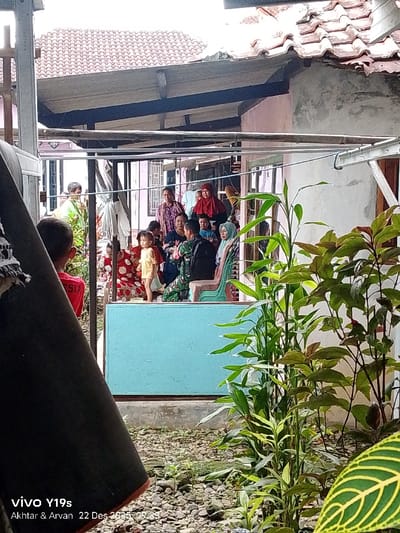 A group of people gathers outside a house. There are several adults and children present, some sitting on chairs, others standing. A young child in a yellow dress stands at the edge of a raised blue platform. The background includes the roof of a building and a partially visible clothesline. Lush greenery obscures parts of the scene. A portion of a mobile phone, its model name and date are visible on the bottom left corner of the image.
