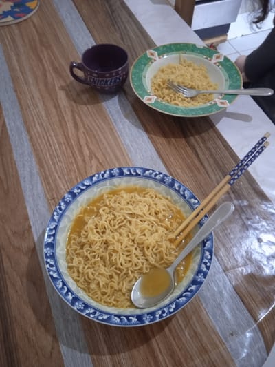 The image shows a wooden table with two bowls of noodles, a purple cup with the word "SNICKERS" on it, a fork, a pair of chopsticks, and a spoon. The noodles are in a clear broth. The location is a home in Réghaïa, Algeria, with the coordinates 36.729, 3.369. The scene is a simple meal, with the bowls of noodles sitting on the table. The spoon is laying across the bowl of noodles, and the chopsticks are lying beside the spoon. The fork is next to the bowl of noodles on the right side of the table. A purple cup with "SNICKERS" printed on it is visible in the top left of the image. There is a partial view of a person sitting in the background. The person is wearing blue jeans and a light-colored shirt. The wooden tabletop has a light brown finish.